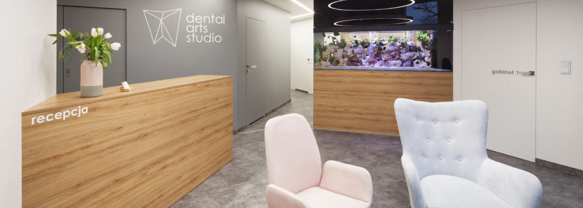 Jasne wnętrze kliniki z recepcją z drewnianym blatem, logo na ścianie, akwarium w tle i pastelowymi fotelami dla pacjentów.