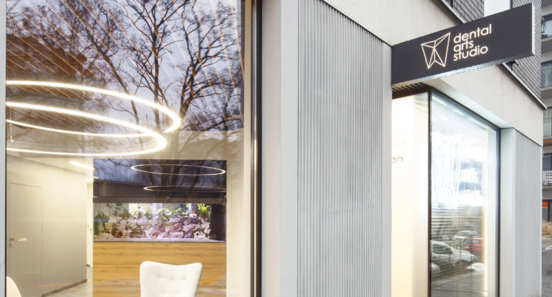 Eleganckie wejście do kliniki z logo 'Dental Arts Studio' na szyldzie, widoczne akwarium wewnątrz i odbicie drzew na szybie.