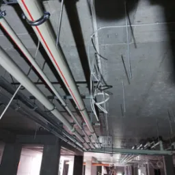 Montaż instalacji hydraulicznej pod stropem w surowym stanie, widoczne rury z oznaczeniami, metalowe uchwyty mocujące i elementy zawieszenia, w tle pracownicy na drabinie.