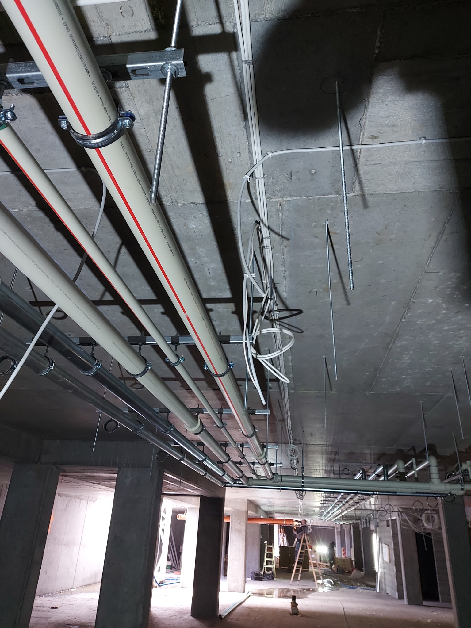 Montaż instalacji hydraulicznej pod stropem w surowym stanie, widoczne rury z oznaczeniami, metalowe uchwyty mocujące i elementy zawieszenia, w tle pracownicy na drabinie.