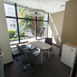 Jasne biuro z owalnym stołem, krzesłami i dużym oknem z widokiem na zieleń. Nowoczesne wnętrze z lampą w kształcie pierścienia i szafką.