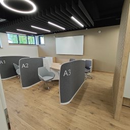 MatchBud Sebastian Zapała - Nowoczesne biuro z drewnianą podłogą i ścianami w odcieniach beżu. Stanowiska pracy oddzielone szarymi panelami z numeracją A1, A2, A3. Biała tablica na ścianie.