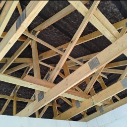 MatchBud Sebastian Zapała - Konstrukcja drewniana dachu z widoczną membraną i elementami łączącymi, wykonana z jasnego drewna, na tle białych ścian. Ujęcie od dołu, ukazujące skomplikowaną geometrię więźby.