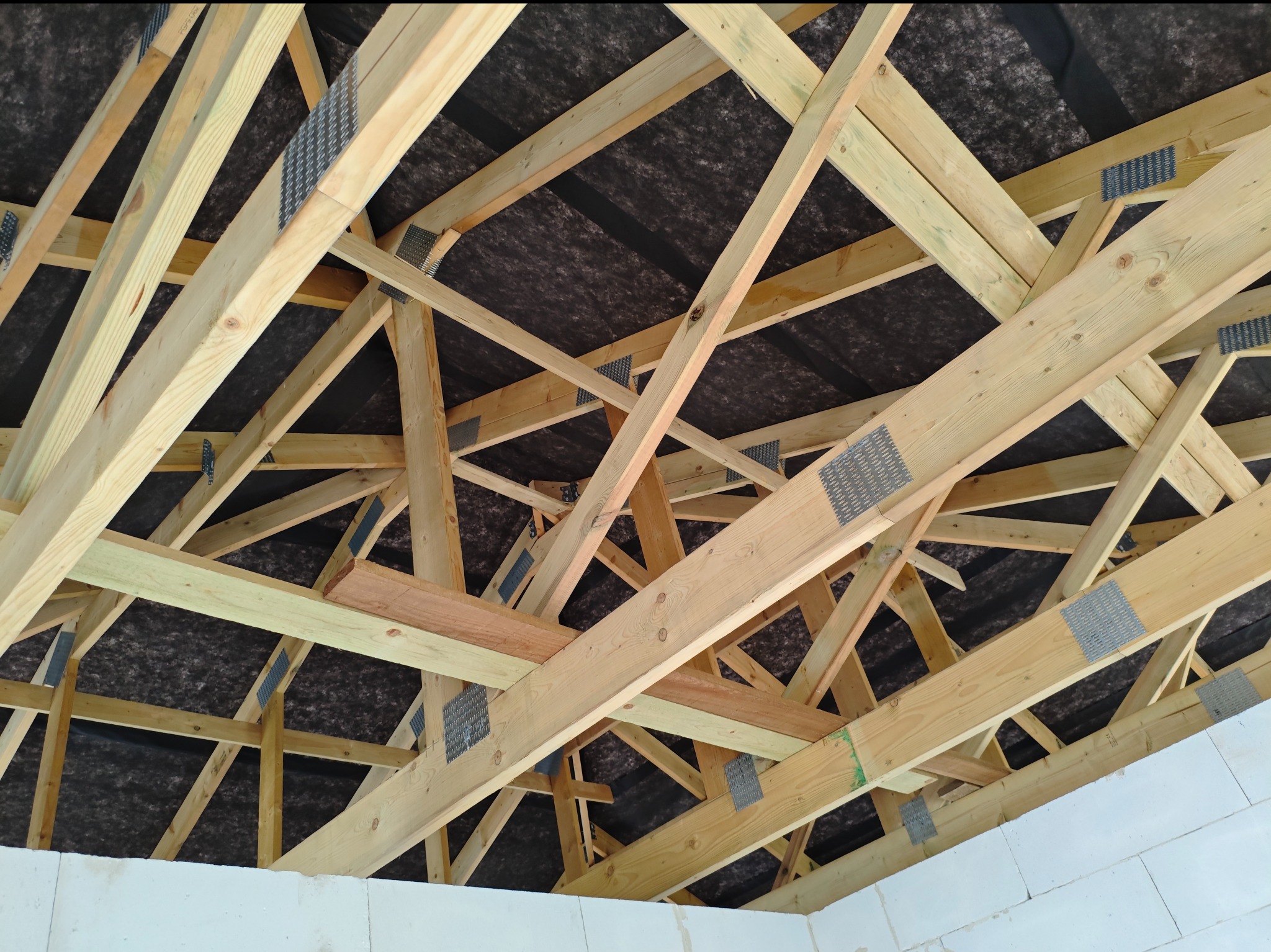 Konstrukcja drewniana dachu z widoczną membraną i elementami łączącymi, wykonana z jasnego drewna, na tle białych ścian. Ujęcie od dołu, ukazujące skomplikowaną geometrię więźby.