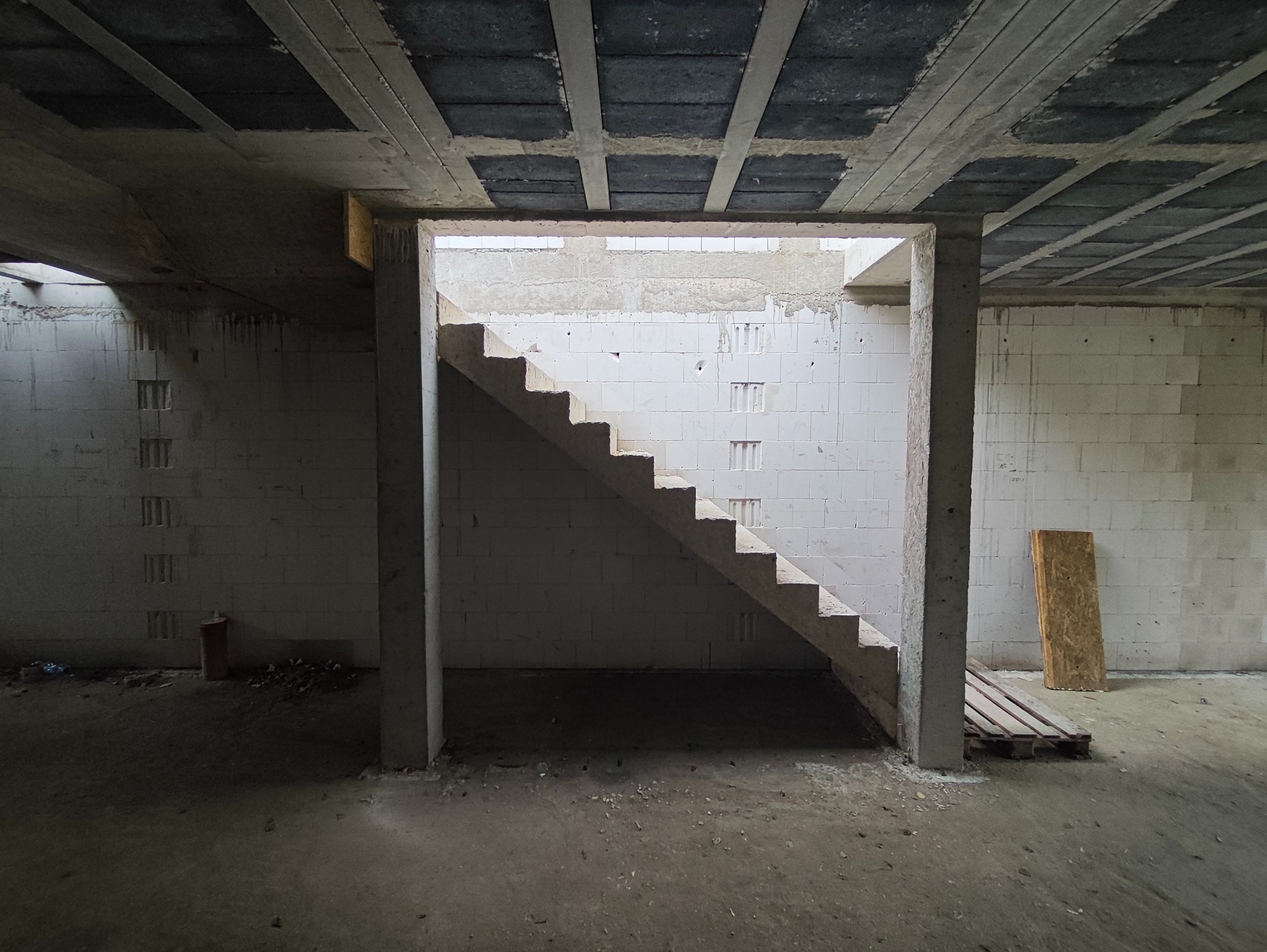 Surowe schody betonowe w trakcie budowy wewnątrz budynku z białymi ścianami z pustaków i betonowym stropem. Widoczne niedokończone elementy i surowy stan wykończenia.