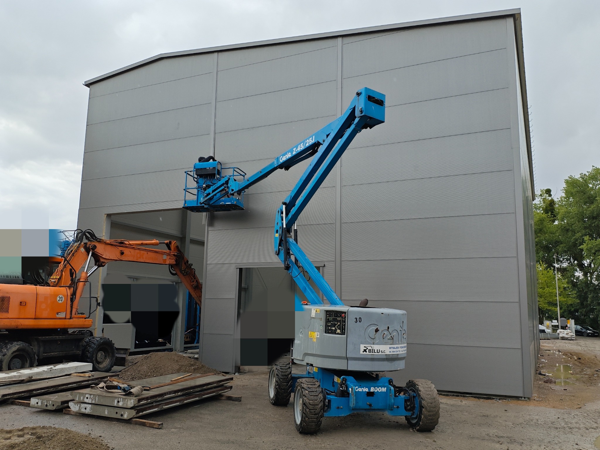 Prace wykończeniowe hali z użyciem podnośnika koszowego Genie Z-45/25J. W tle widoczny pomarańczowy koparka i płyty betonowe. Szara fasada hali z poziomymi panelami.