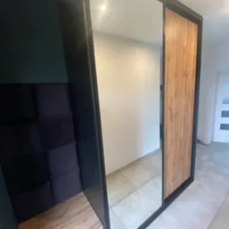 Nowoczesna szafa z lustrzanymi drzwiami przesuwnymi i siedziskiem z tapicerowanym oparciem w ciemnych odcieniach, wkomponowana we wnękę, z widoczną teksturą drewna na jednym z paneli.