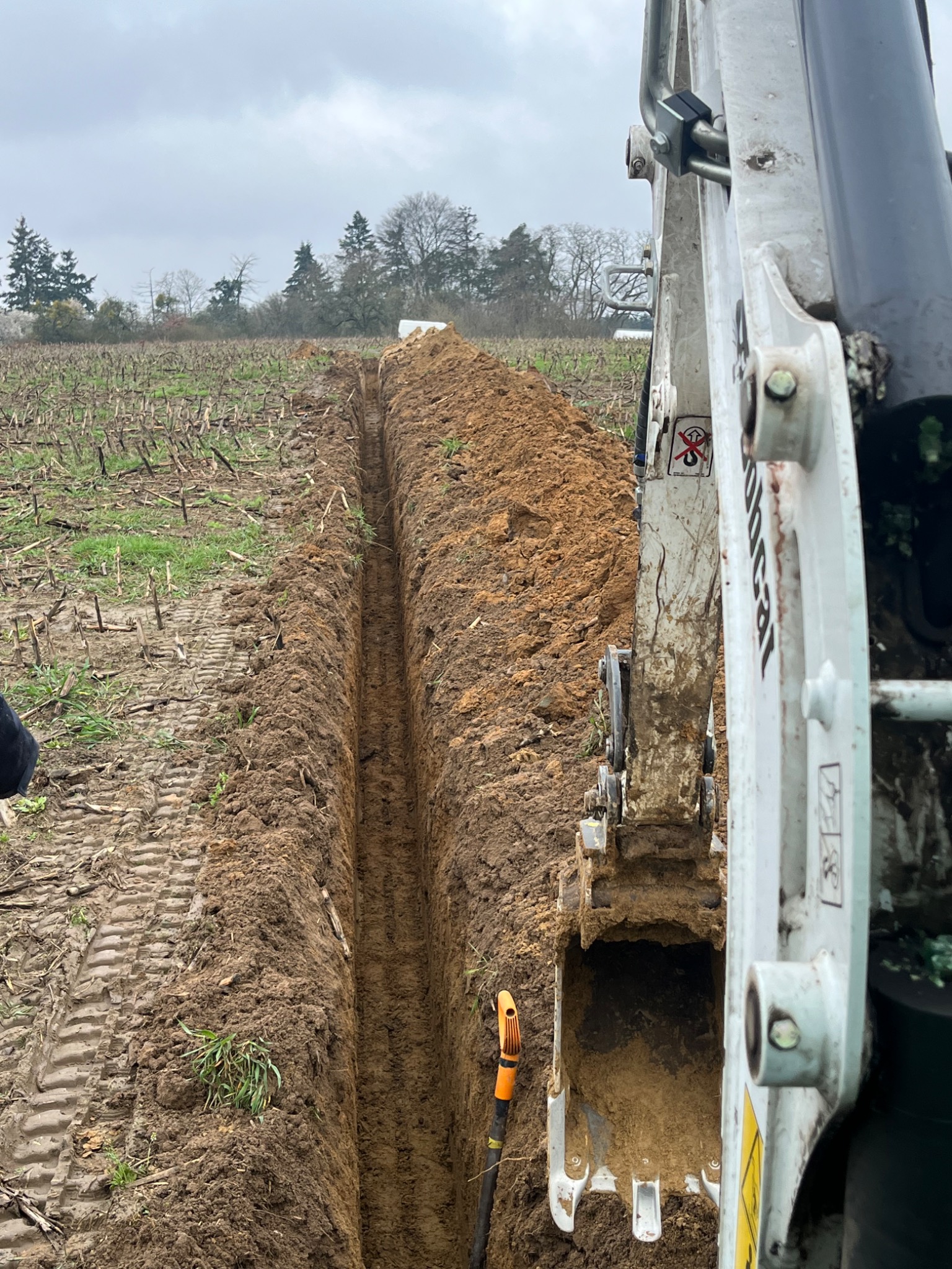 Wąski wykop w polu uprawnym, wykonany przy użyciu minikoparki Bobcat, widoczny pomarańczowy uchwyt narzędzia w wykopie.