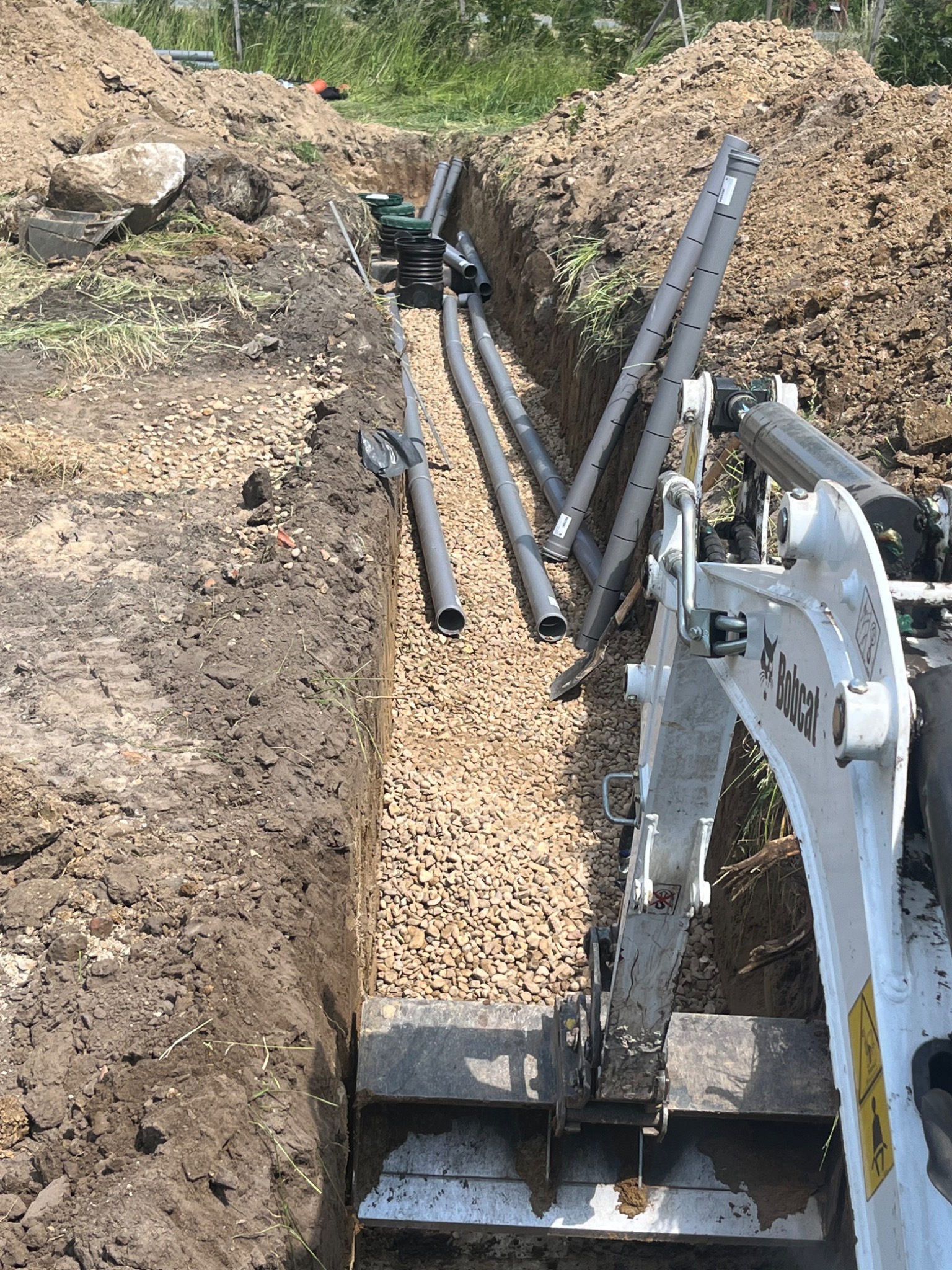 Wykopany rów wypełniony żwirem z ułożonymi rurami drenażowymi, widoczny mini-koparka Bobcat, ziemia po bokach wykopu.
