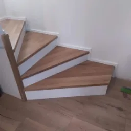 Kątowe ujęcie drewnianych stopni z białymi podstopnicami, prowadzących w górę, z widoczną podłogą z paneli w odcieniach brązu i fragmentem białej ściany z włącznikami światła.