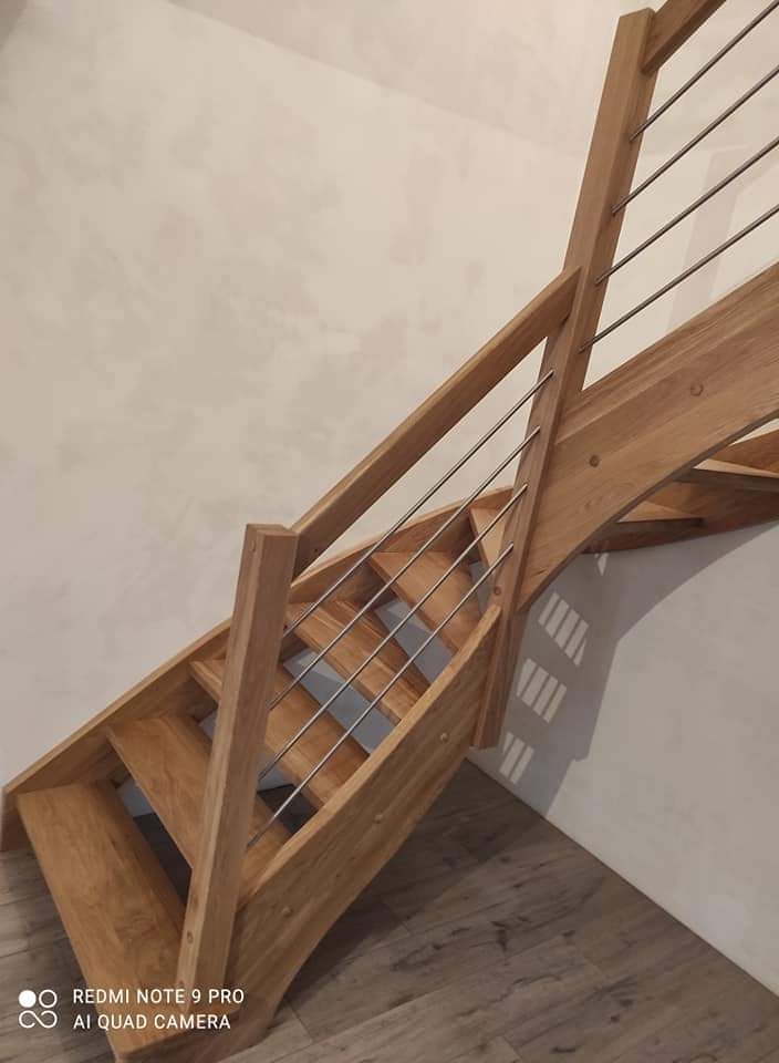 Jasne, dębowe stopnie z metalowymi prętami balustrady, widok od dołu, z widocznym zaokrągleniem konstrukcji.