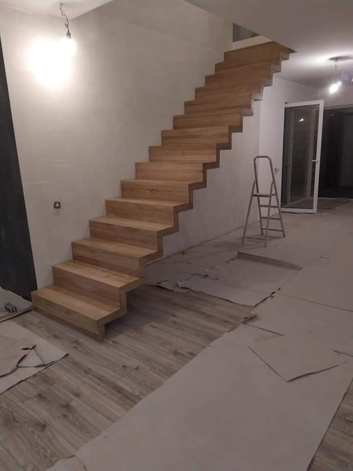 Jasne, drewniane stopnie schodów bez poręczy, zamontowane na betonowej konstrukcji, widoczne w trakcie prac wykończeniowych, zabezpieczone kartonem podłogi, drabina oparta o ścianę.