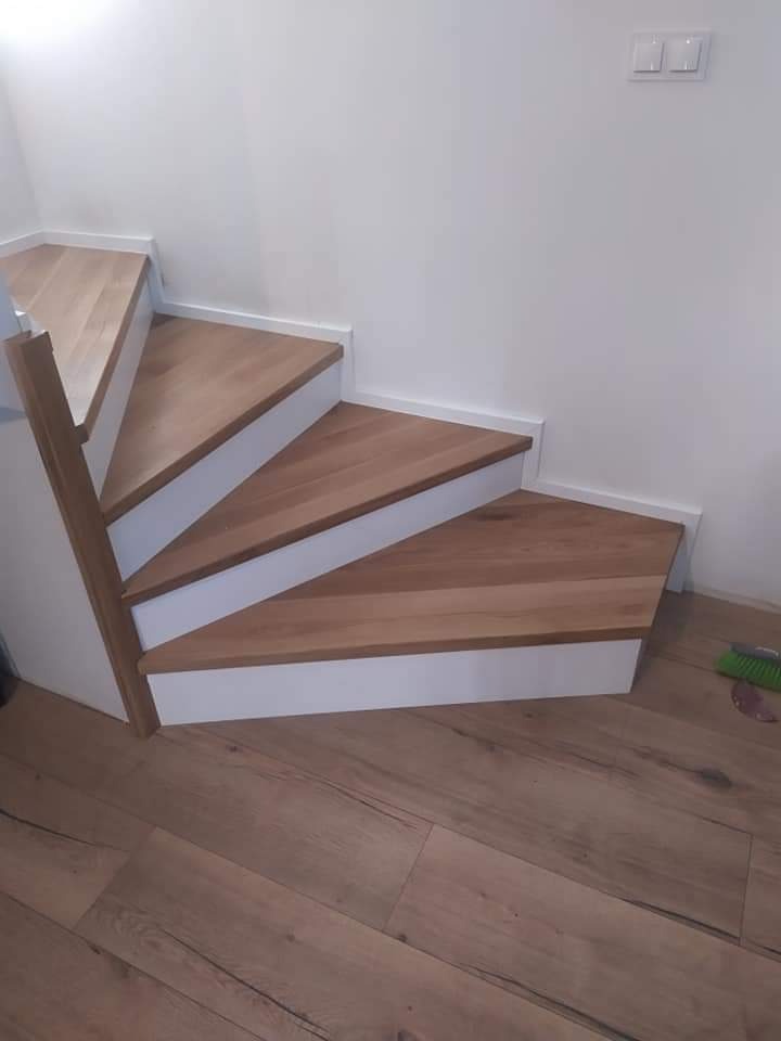 Kątowe ujęcie drewnianych stopni z białymi podstopnicami, prowadzących w górę, z widoczną podłogą z paneli w odcieniach brązu i fragmentem białej ściany z włącznikami światła.