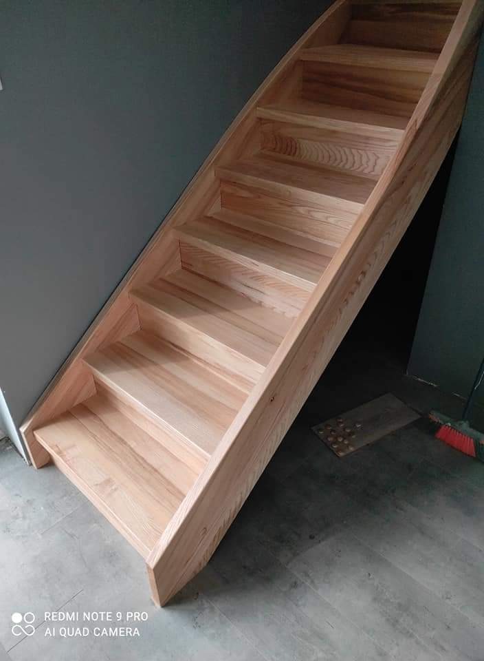 Jasne, drewniane schody bez poręczy, ustawione pod kątem, z widocznym usłojeniem drewna, na tle szarej ściany i podłogi, z fragmentem listwy przypodłogowej i szczotki widocznymi w dolnym rogu.