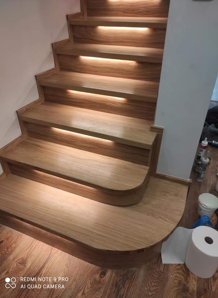 Drewniane schody z podświetleniem LED pod stopniami, widoczne materiały budowlane obok ściany, parkiet drewniany.