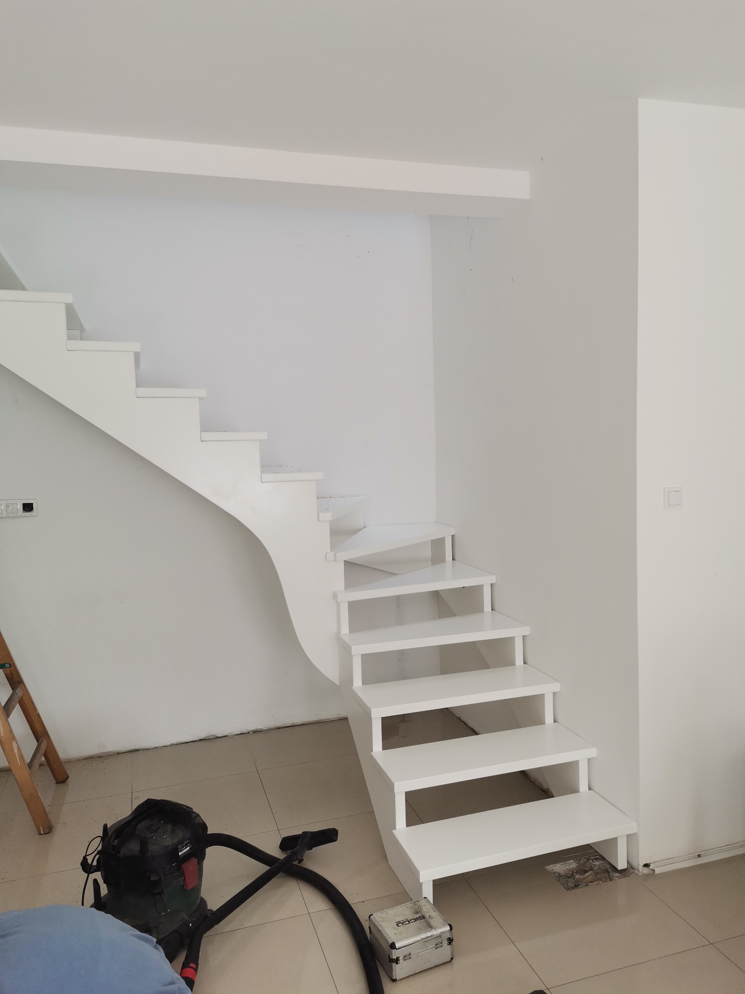 Białe, nowoczesne schody o nietypowym kształcie z zakrzywionym bokiem, widoczne podczas montażu, z odkurzaczem przemysłowym i narzędziami obok.