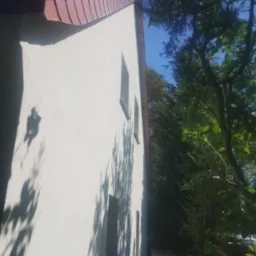 Boczny widok otynkowanego domu z dwoma oknami i czerwoną dachówką, z widocznymi cieniami drzew na elewacji i bujną zielenią w tle.