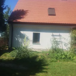 Boczny widok domu z jasną elewacją, brązową dachówką karpiówką z oknem dachowym i kominem, zarośnięty ogród z drabinką dla pnączy przy ścianie.