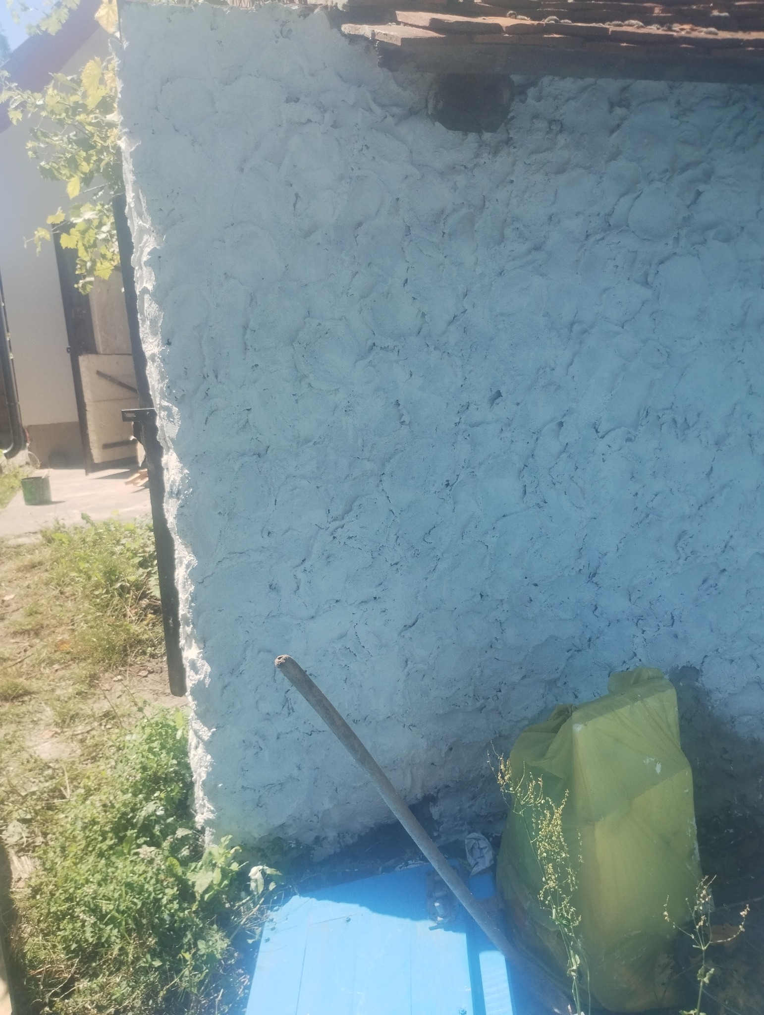 Fragment ściany budynku z nierówną, białą strukturą tynku, widoczny z poziomu gruntu, z elementami roślinności i narzędziem opartym o ścianę, obok leży worek na odpady.