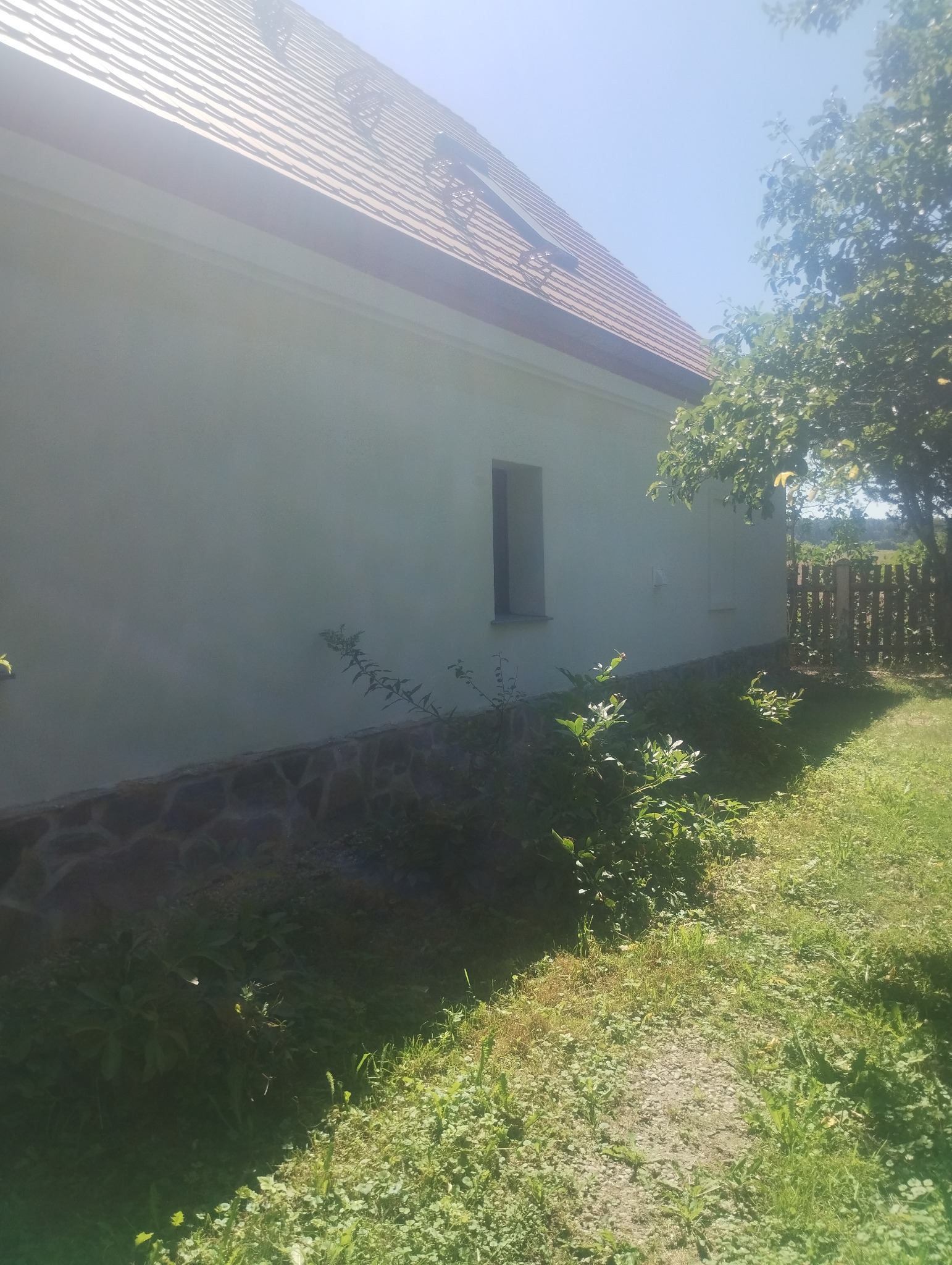 Boczny widok otynkowanego domu jednorodzinnego z niewielkim oknem, widoczne krzewy i fragment kamiennej podmurówki, w tle drewniany płot i zieleń.