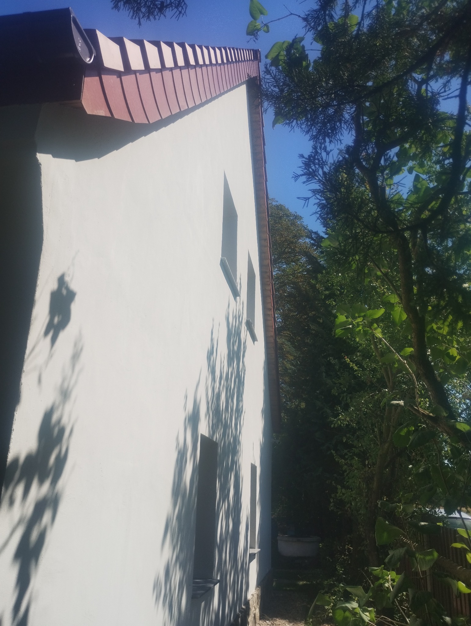 Boczny widok otynkowanego domu z dwoma oknami i czerwoną dachówką, z widocznymi cieniami drzew na elewacji i bujną zielenią w tle.