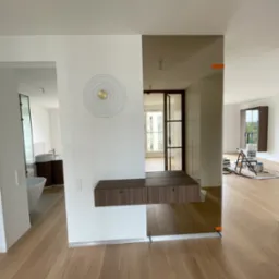 Nowoczesne wnętrze z drewnianą konsolą wiszącą na ścianie, lustrem dzielącym przestrzeń i widokiem na łazienkę oraz pokój w trakcie malowania.