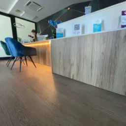 Recepcja z blatem drewnianym o wyraźnym usłojeniu, podświetlona od spodu, z dwoma krzesłami w odcieniach zieleni i błękitu, widok z niskiej perspektywy na podłogę.