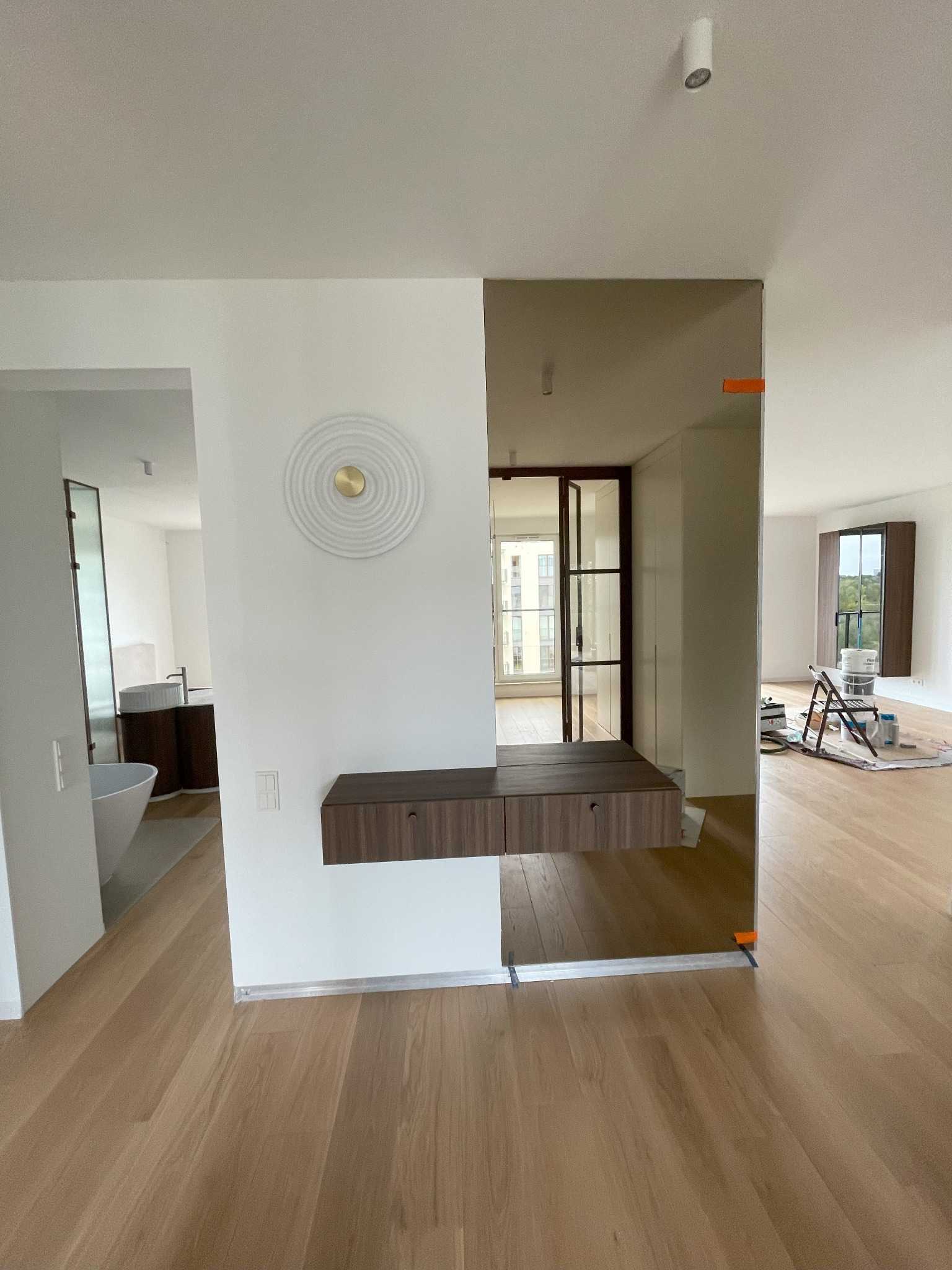 Nowoczesne wnętrze z drewnianą konsolą wiszącą na ścianie, lustrem dzielącym przestrzeń i widokiem na łazienkę oraz pokój w trakcie malowania.