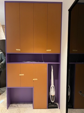 Pomarańczowo-fioletowa szafa na wymiar z półką i miejscem na odkurzacz pionowy, widoczny fragment osoby po lewej stronie.