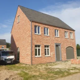 Nowoczesny, dwukondygnacyjny dom murowany z czerwonej cegły z szarą dach&oacute;wką, białymi oknami i szarymi drzwiami garażowymi; na pierwszym planie zaparkowany biały samoch&oacute;d dostawczy budowlanej firmy...