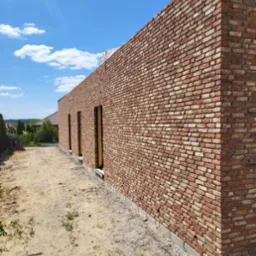 Wysoka ściana z czerwonej cegły z trzema otworami okiennymi bez okien, w trakcie budowy, widok z boku, na piasku z zielenią, w tle zabudowania i niebo z chmurami.