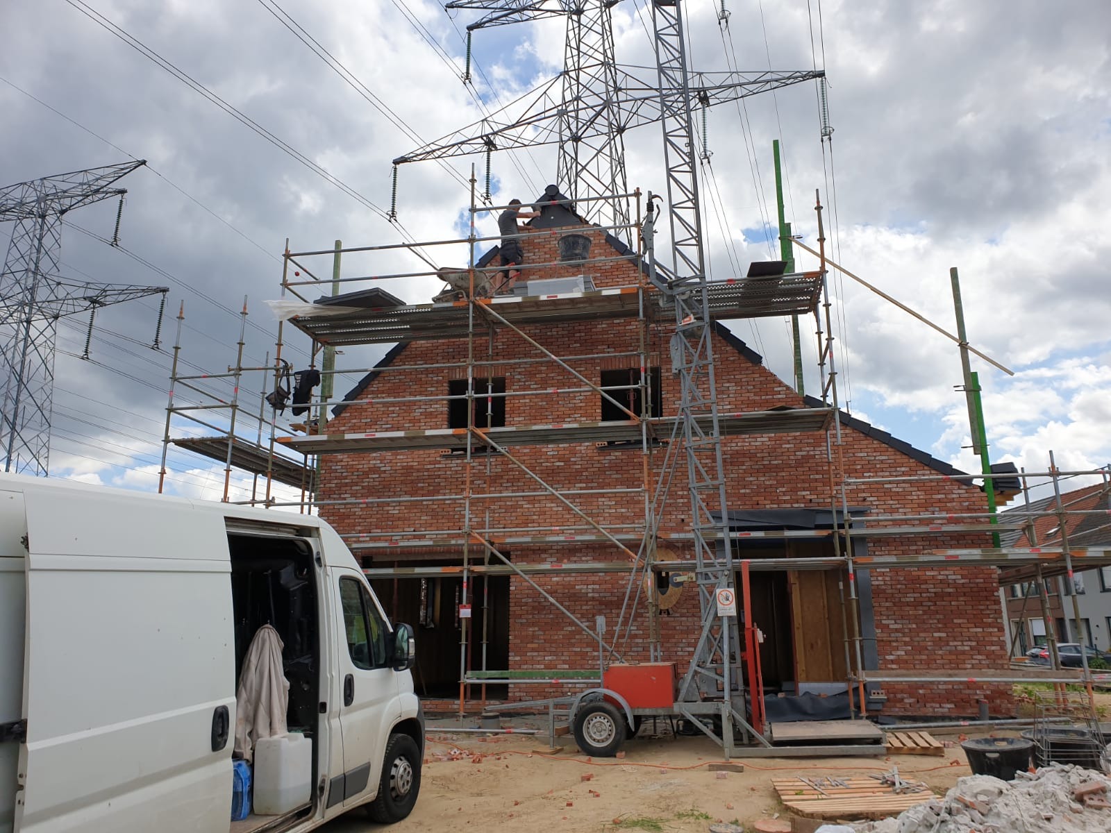 Budowa domu z czerwonej cegły na etapie murowania ścian zewnętrznych, widoczne rusztowania, pracownicy na dachu i linia wysokiego napięcia w tle.