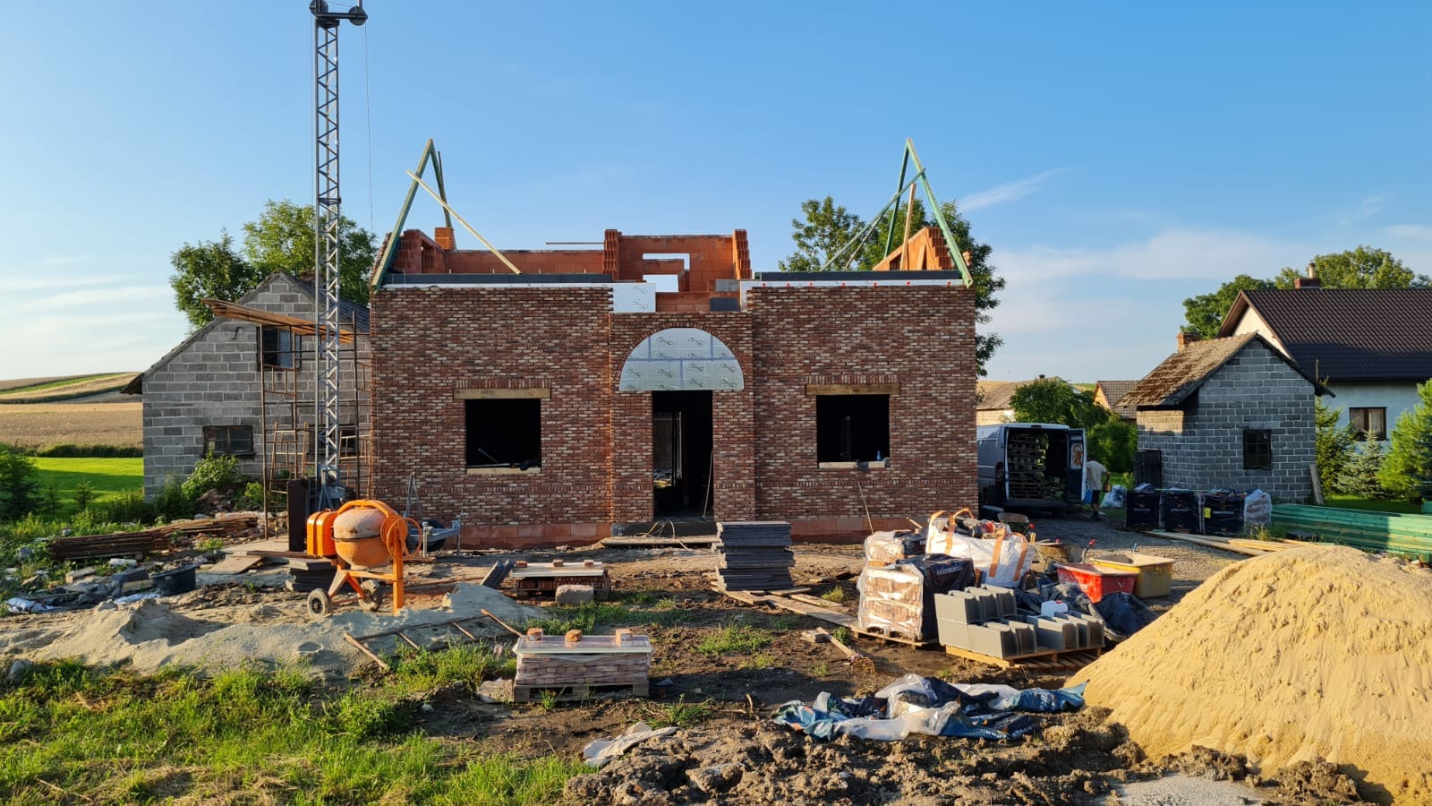 Wznoszenie ścian z czerwonej cegły klinkierowej w trakcie budowy domu, widoczna betoniarka, rusztowania i sterty materiałów budowlanych.