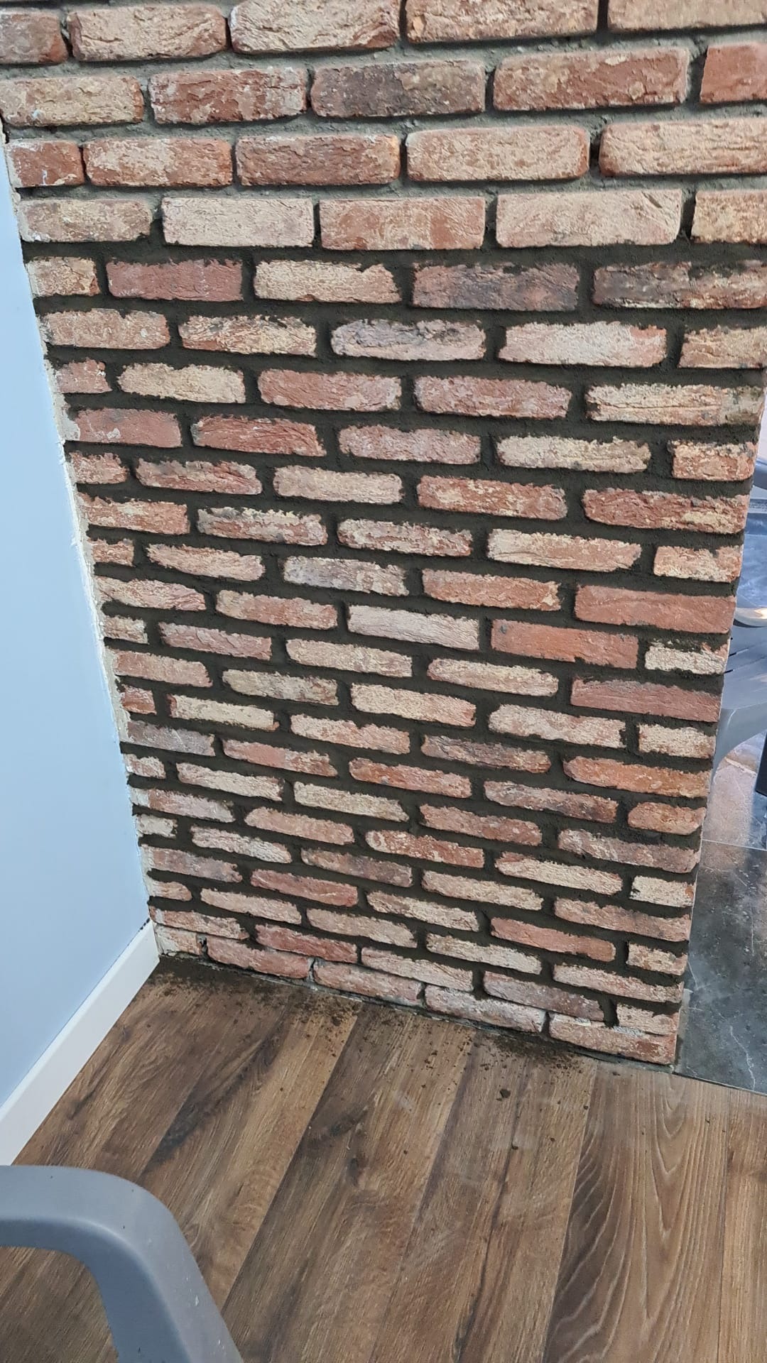 Wnętrze z fragmentem ściany wykończonej cegłą klinkierową w odcieniach czerwieni i beżu, z widoczną fugą w kolorze ciemnoszarym, na tle drewnianej podłogi i błękitnej ściany.