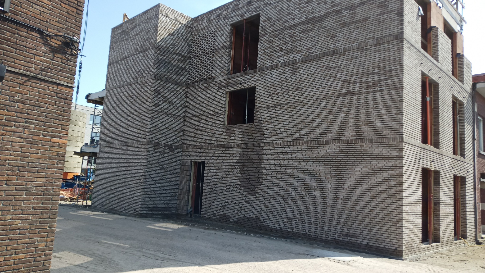 Surowy stan murowanego budynku z szarej cegły z otworami okiennymi i drzwiowymi, widoczne rusztowania w tle.