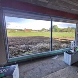 Duże okno aluminiowe z widokiem na pole uprawne, w trakcie montażu, widoczne narzędzia i materiały budowlane w surowym wnętrzu.