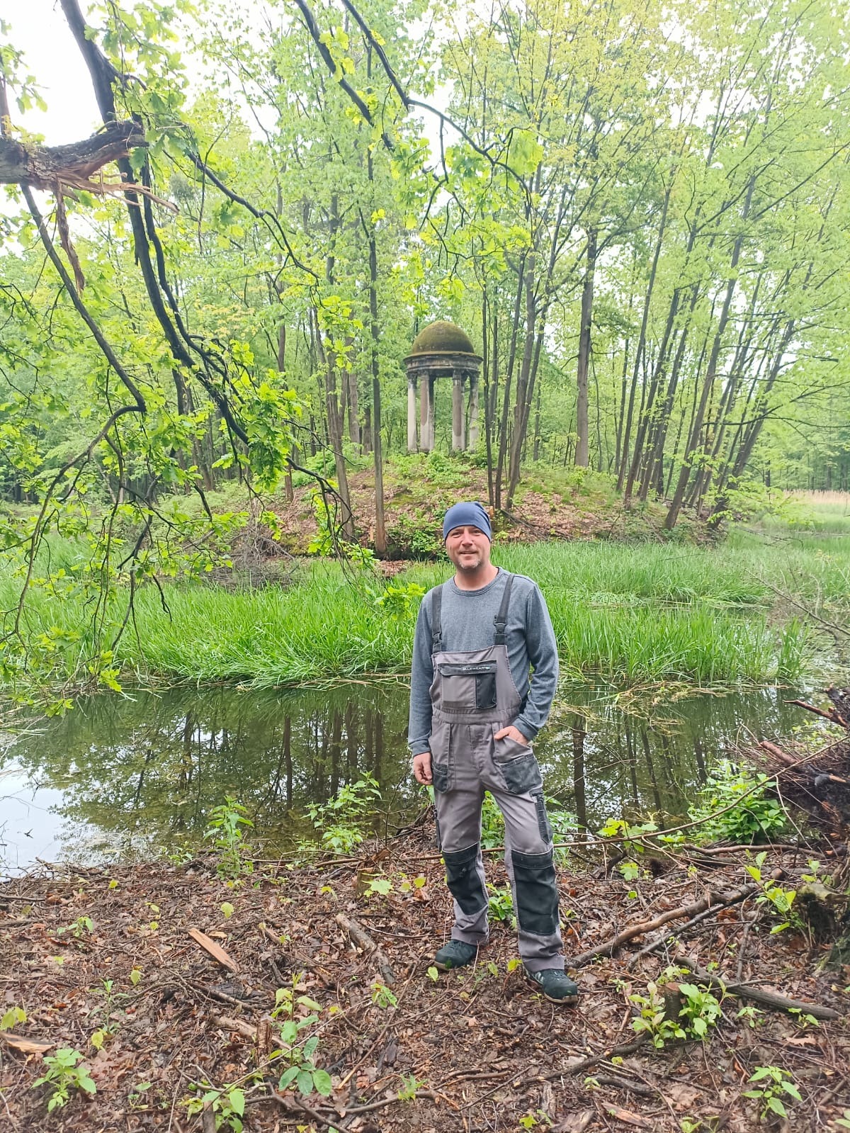 Pracownik w szarym kombinezonie roboczym stoi nad brzegiem stawu we Wrocławiu, w tle altana w parku.