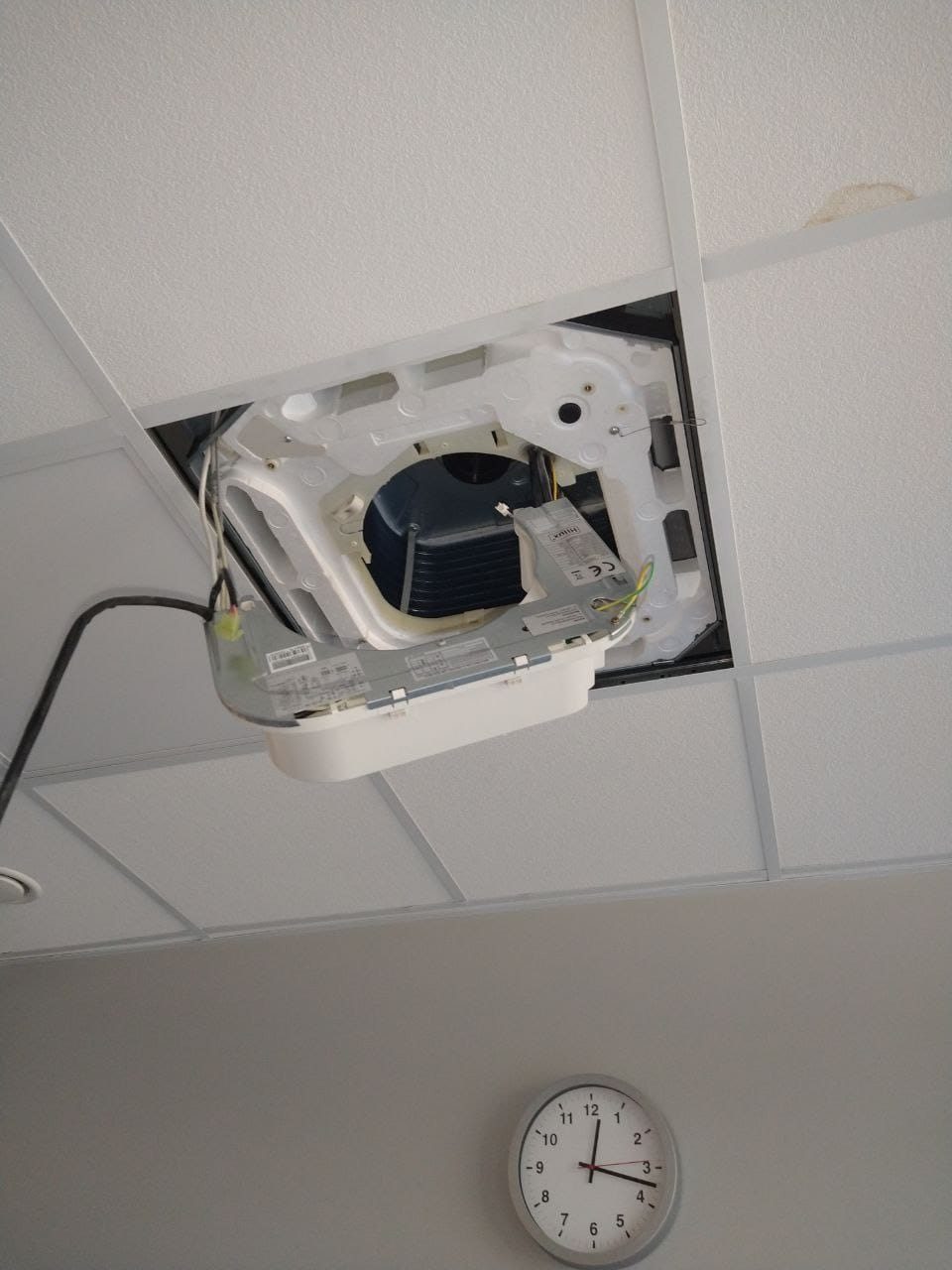 Otwarta obudowa sufitowa klimatyzacji w kasetonowym suficie, widoczne elementy wewnętrzne i przewody, zegar na ścianie poniżej.