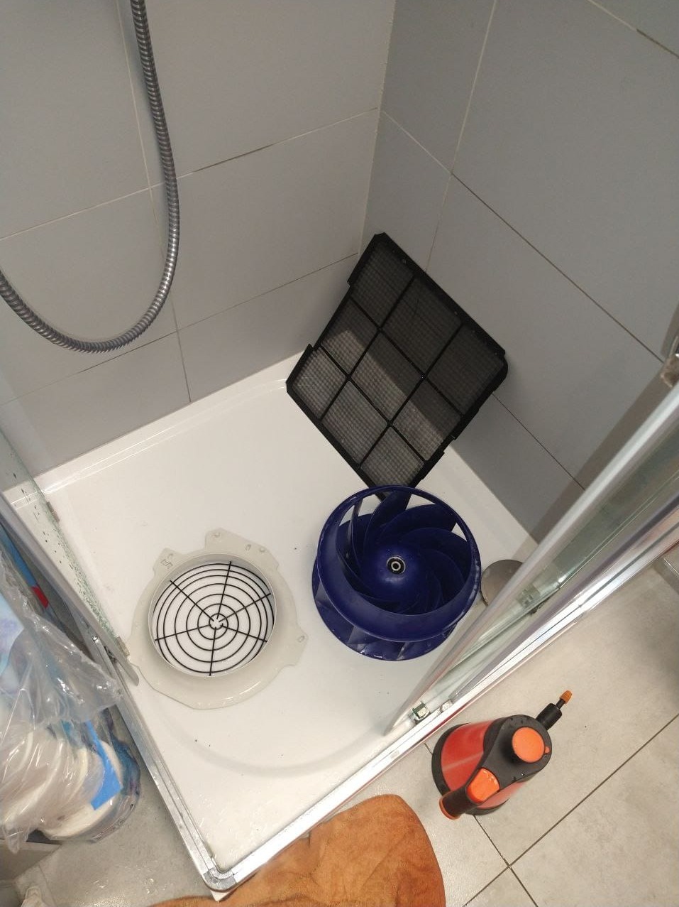 Elementy wentylacji łazienkowej: filtr kratowy, wirnik wentylatora w kolorze niebieskim oraz kratka zabezpieczająca, leżące w brodziku prysznicowym podczas czyszczenia.