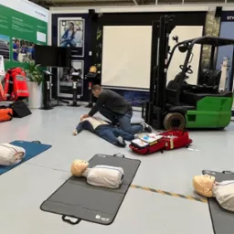 Instruktor demonstruje resuscytację krążeniowo-oddechową na manekinie podczas szkolenia w Katowicach. W tle wózek widłowy i materiały informacyjne.