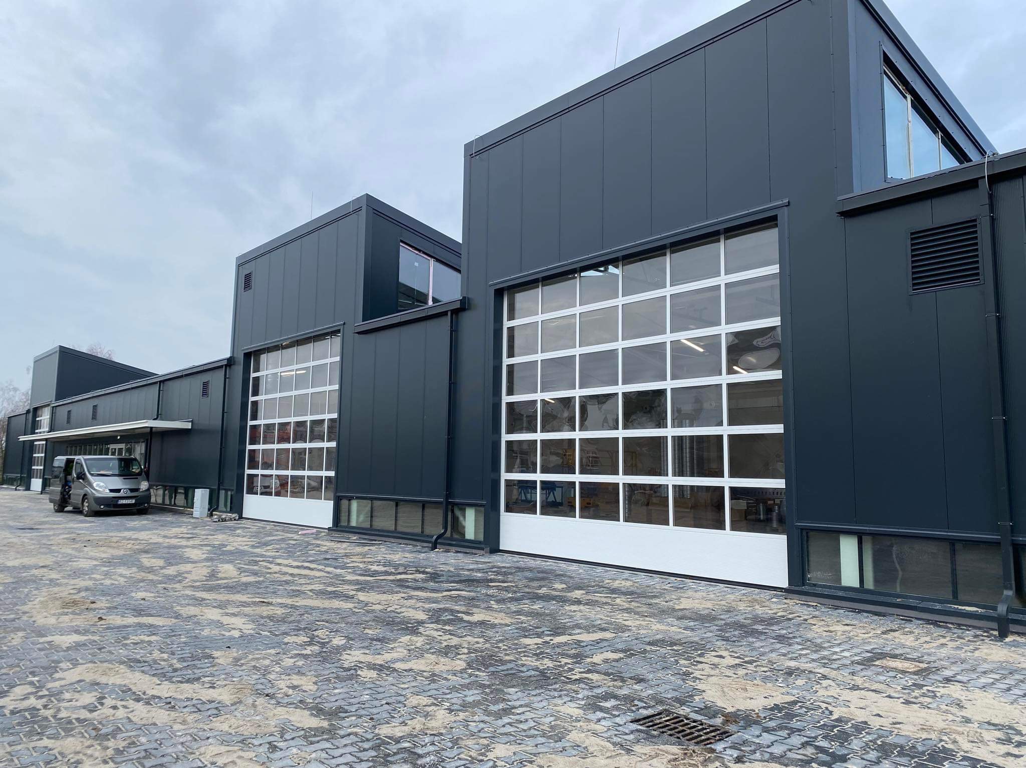 Nowoczesna hala przemysłowa z szarymi ścianami z płyt i segmentowymi bramami garażowymi z białymi ramami, widok z zewnątrz na kostkę brukową i zaparkowany szary samochód dostawczy.