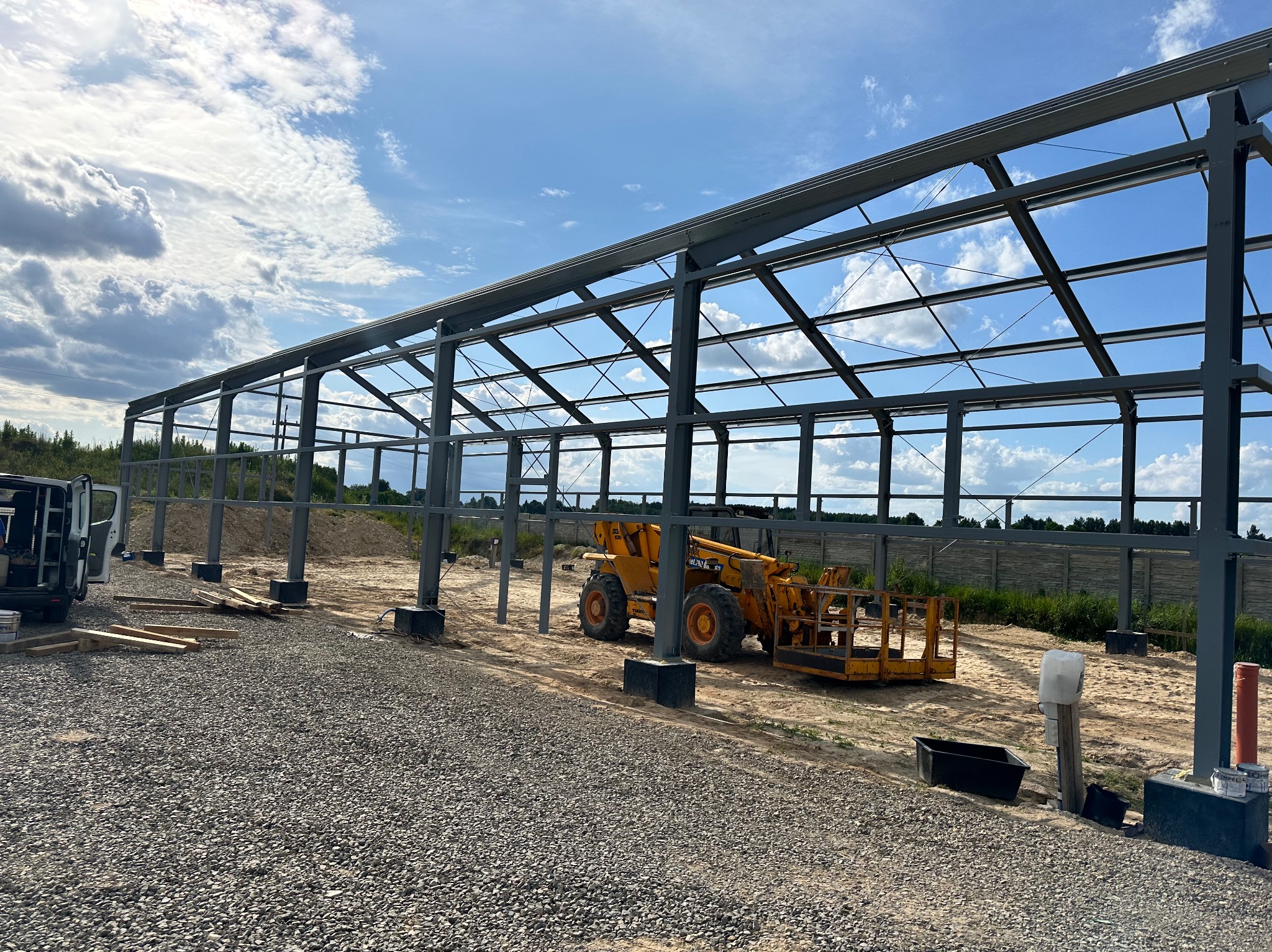 Stalowa konstrukcja hali w trakcie budowy, żółta ładowarka teleskopowa na piaszczystym podłożu, widoczne fundamenty i niebo z chmurami.