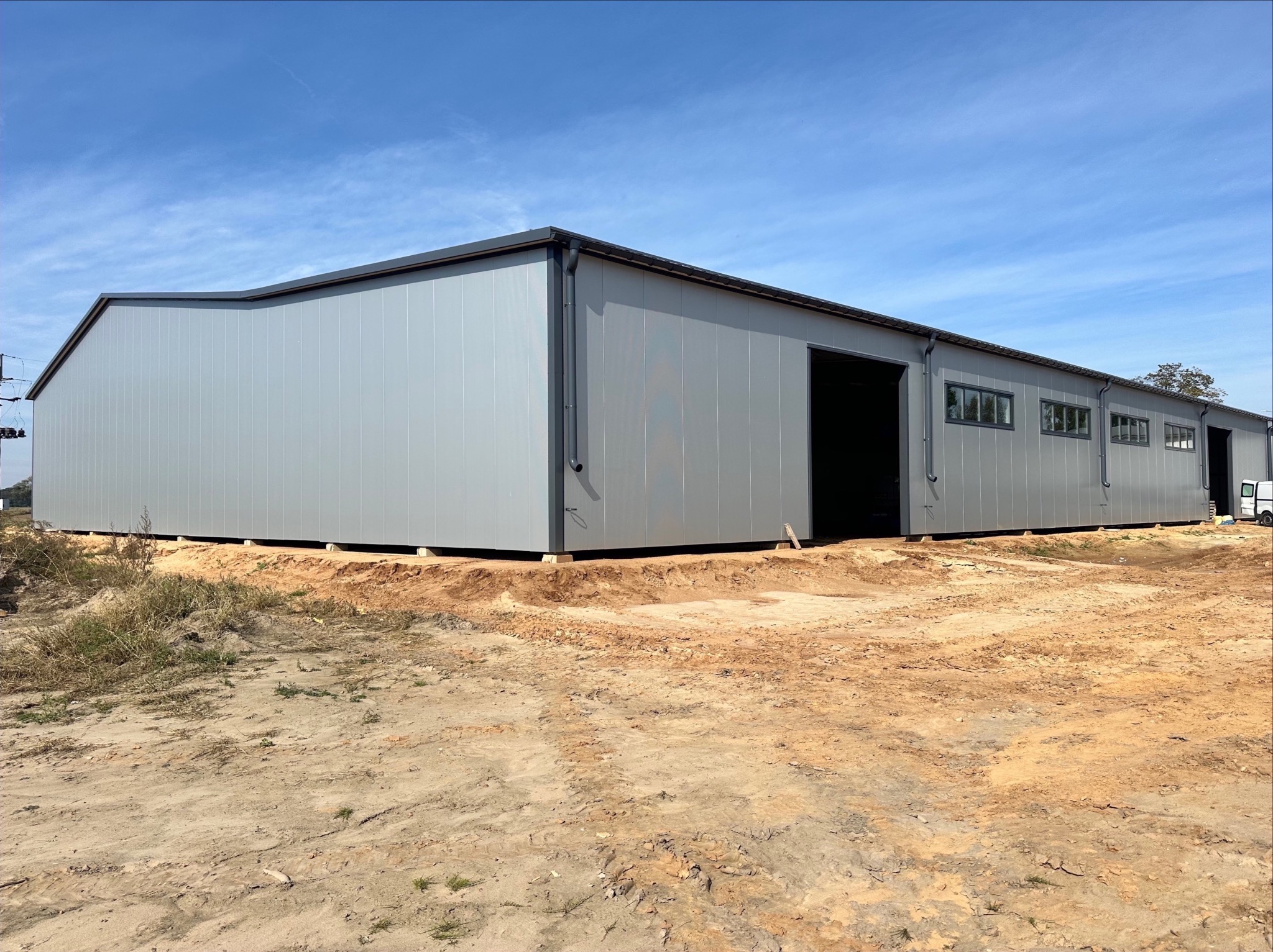 Nowoczesna hala przemysłowa z szarych płyt warstwowych, z widocznymi oknami i otwartymi wrotami, posadowiona na betonowych fundamentach, otoczona ziemią i skąpą roślinnością, pod błękitnym niebem.