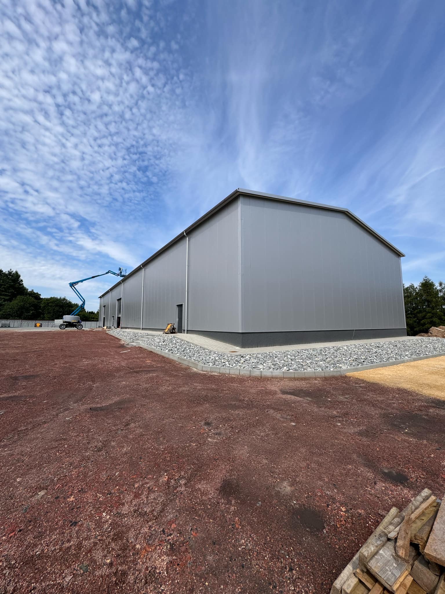 Nowo wybudowana hala z szarych płyt warstwowych, z widocznym podnośnikiem koszowym w trakcie prac wykończeniowych i przygotowanym terenem wokół budynku.