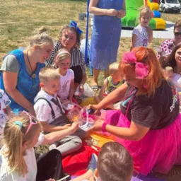 Animator w różowej spódnicy rozdaje dzieciom kolorowe kredki na kocu piknikowym, w tle dmuchany zamek i osoby dorosłe.