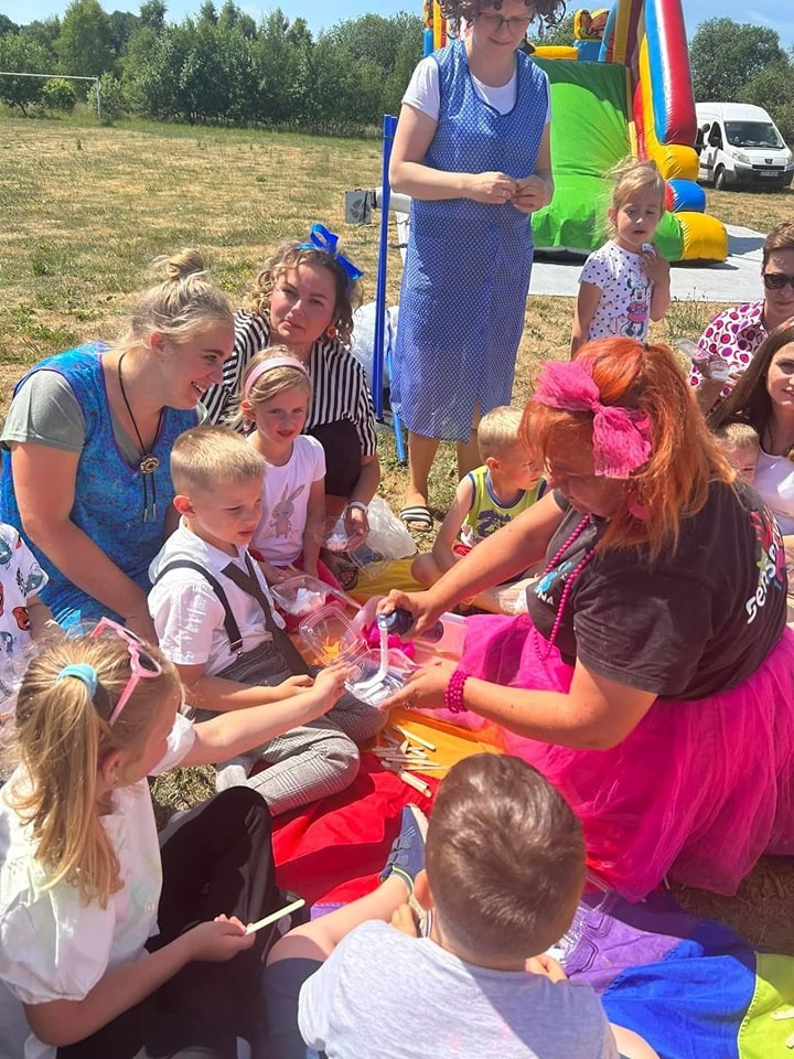 Animator w różowej spódnicy rozdaje dzieciom kolorowe kredki na kocu piknikowym, w tle dmuchany zamek i osoby dorosłe.