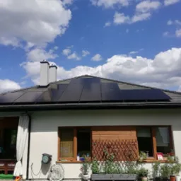 Czarny panel słoneczny na dachu domu jednorodzinnego z szarą dachówką, na tle błękitnego nieba z białymi chmurami. Widoczny fragment białej elewacji z oknem i roślinami w doniczkach.