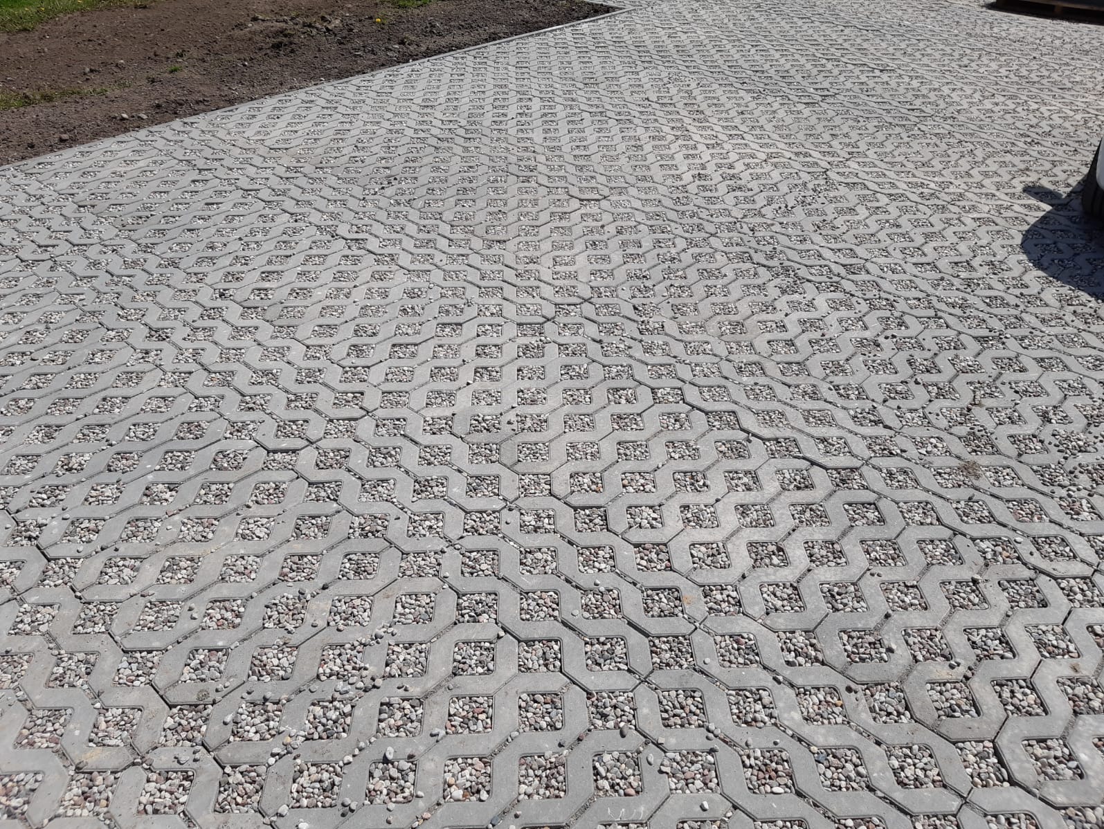 Szara, betonowa ażurowa nawierzchnia z wypełnieniem żwirowym, ułożona na posesji. Widoczny fragment samochodu w prawym górnym rogu kadru.