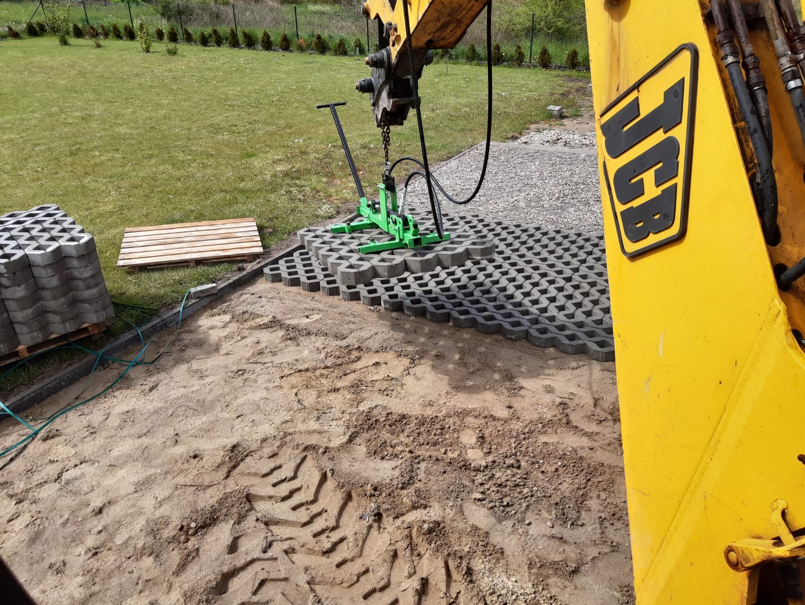 Układanie ażurowych płyt betonowych na piasku za pomocą chwytaka podwieszonego do koparki JCB. Widoczne ślady opon i paleta z kostką.