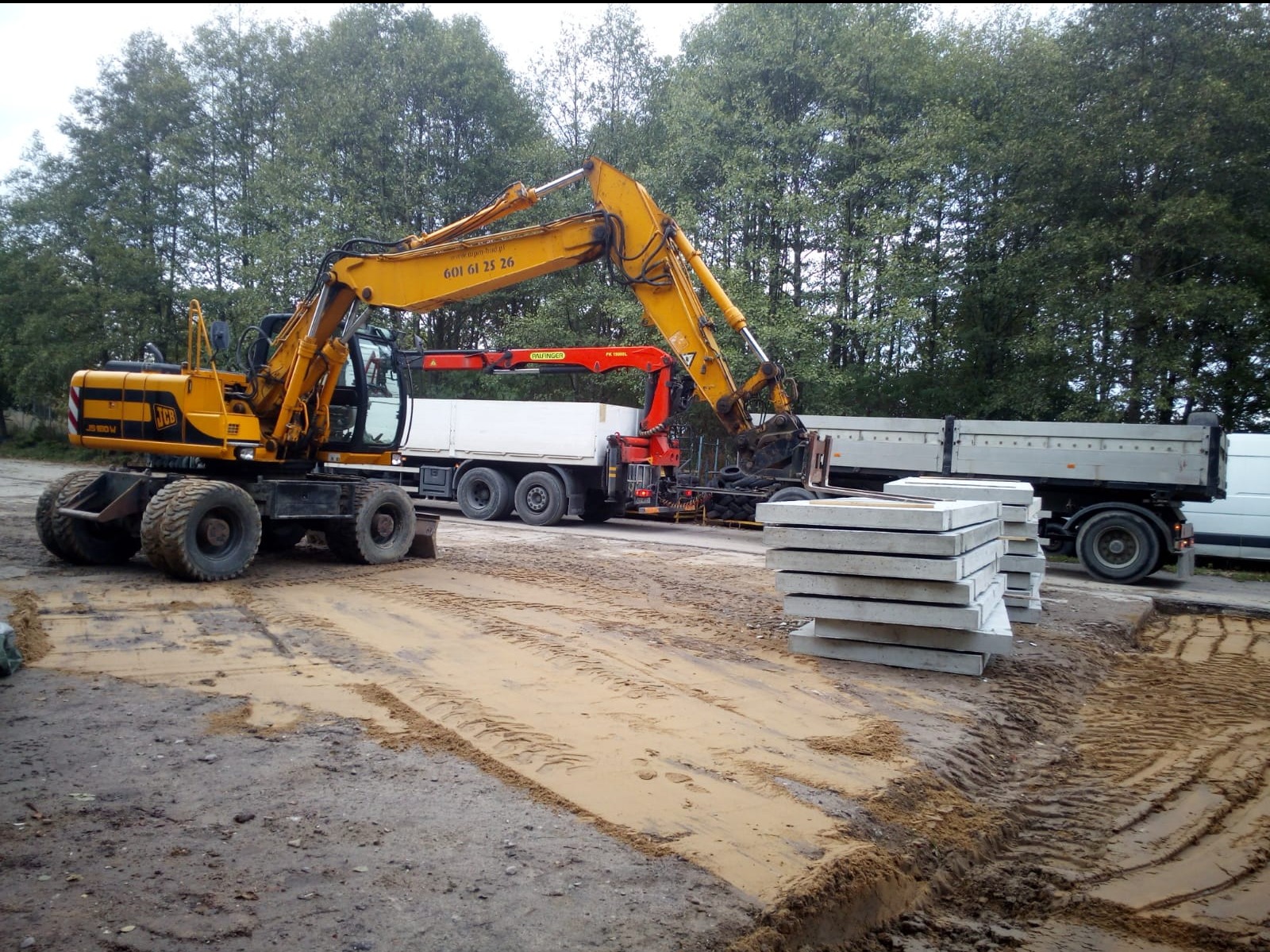 Żółta koparka JCB na kołach, obok ciężarówka z dźwigiem i paleta betonowych płyt. Przygotowany teren pod budowę, widoczne ślady opon w błocie.